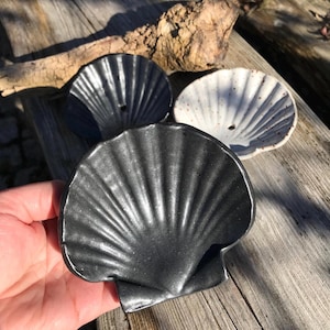 Könnte beinhalten: Drei keramische Schalen in Form von Muscheln. Eine ist schwarz, eine weitere gesprenkelt weiß und die dritte dunkelblau. Die Schalen haben eine gerippte Textur und ein kleines Loch in der Mitte. Sie werden auf einer verwitterten Holzoberfläche präsentiert.
