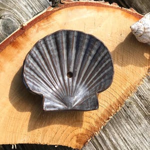 Könnte beinhalten: Eine seeförmige Seifenschale aus Keramik mit dunkelblauer und weißer Glasur. Die Schale hat ein kleines Loch in der Mitte und steht auf einer Holzoberfläche. Im Hintergrund ist eine Muschel zu sehen.