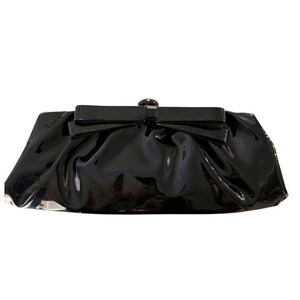 May include: A glossy black clutch with a bow-shaped clasp and a small, round, dark brown button. The clutch has a gathered design and a shiny, patent leather-like finish. The bag is rectangular and appears to be empty.