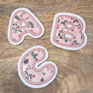 Könnte beinhalten: Drei Filzbuchstaben, A, B und C, in einem rosa Blumenstoff mit weißen Nähten. Die Buchstaben befinden sich auf einer Holzoberfläche.