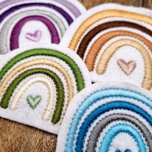 Könnte beinhalten: Fünf gestickte Filz-Regenbogen-Patches mit einem Herz in der Mitte jedes. Die Regenbögen sind in verschiedenen Farbkombinationen, darunter Lila, Grün, Braun und Blau.