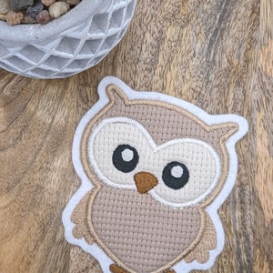Könnte beinhalten: Eine braune gestickte Eulen-Applikation mit weißen Nähten und schwarzen Augen. Die Eule befindet sich auf einer Holzoberfläche.