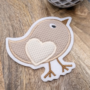 Könnte beinhalten: Ein Stoffaufnäher in Form eines Vogels. Der Vogel ist hauptsächlich beige mit einem herzförmigen Design auf der Brust. Er hat einen weißen Rand, ein schwarzes Auge und braune Füße und einen Schnabel. Der Aufnäher ist zum Aufnähen oder Aufbügeln auf Stoff gedacht.