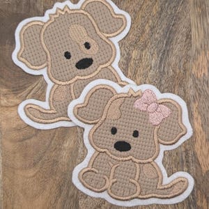 Könnte beinhalten: Zwei gestickte Aufnäher mit einem Cartoon-Welpen-Design. Die Welpen sind hellbraun mit dunkelbraunen Akzenten und schwarzen Augen. Ein Welpe hat eine rosa Schleife. Die Aufnäher haben einen weißen Rand und liegen auf einer Holzoberfläche.