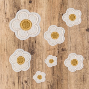 Könnte beinhalten: Eine Sammlung weißer Gänseblümchen-Blumenaufnäher mit gelben Zentren und cremefarbenen Stickereidetails. Die Aufnäher sind unterschiedlich groß und auf einer Holzoberfläche angeordnet.