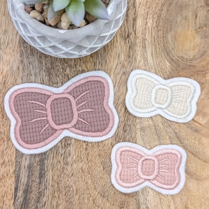 Könnte beinhalten: Drei gestickte Stoff-Schleifen-Patches in verschiedenen Größen. Die größte Schleife ist rosa, die mittlere Schleife ist cremefarben und die kleinste Schleife ist rosa.