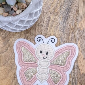 Könnte beinhalten: Eine rosa und goldene Glitzer-Schmetterlings-Applikation auf weißem Hintergrund mit einem lächelnden Gesicht. Der Schmetterling ist mit einem detaillierten Design bestickt.