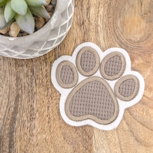 Könnte beinhalten: Ein gestickter Pfotenabdruck-Aufnäher mit weißem Rand und braunen Details. Der Pfotenballen und die Zehen sind strukturiert braun. Der Aufnäher liegt auf einer Holzoberfläche, mit einem dekorativen Topf und einer Sukkulentenpflanze im Hintergrund.