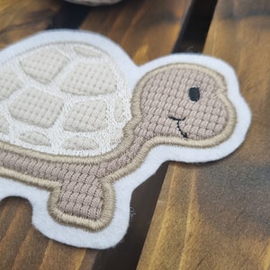 Könnte beinhalten: Eine weiße Filzapplikation einer braunen Schildkröte mit einem gestickten Panzerdesign. Die Schildkröte hat ein lächelndes Gesicht mit einem schwarzen Auge.