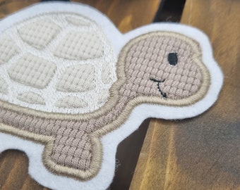 May include: A white felt applique of a brown turtle with a stitched shell design. The turtle has a smiling face with a black eye.