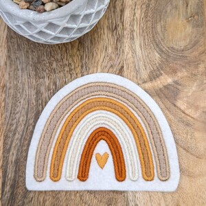 Könnte beinhalten: Ein weißer Filz-Patch mit einem gestickten Regenbogen in Braun-, Orange- und Cremetönen. Ein kleines Herz ist in der Mitte des Regenbogens gestickt.