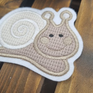 Könnte beinhalten: Eine beige gestickte Schnecken-Applikation mit weißem Umriss. Die Schnecke hat eine braune Schale und einen braunen Körper mit schwarzen Augen.