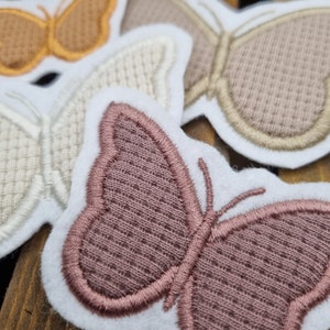 Könnte beinhalten: Vier gestickte Schmetterlings-Patches in verschiedenen Farben und Texturen. Die Patches sind weiß mit einem braunen, beige und rosa Schmetterlingsdesign.