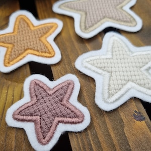 Könnte beinhalten: Vier gestickte Filzsternformen in verschiedenen Farben: Beige, Braun, Rosa und Weiß. Die Sterne liegen auf einer Holzoberfläche.