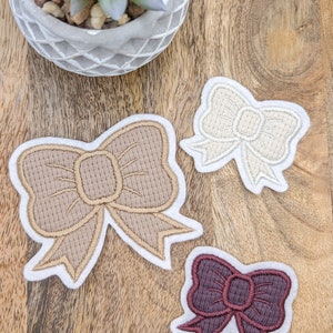 Könnte beinhalten: Drei gestickte Stoff-Schleifen-Patches in verschiedenen Größen und Farben. Die größte Schleife ist beige, die mittlere Schleife ist weiß und die kleinste Schleife ist burgunderrot.