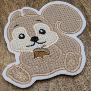 Könnte beinhalten: Gestickter Aufnäher mit einem Cartoon-Eichhörnchen, das eine Eichel hält. Das Eichhörnchen hat einen hellbraunen Körper mit einem strukturierten Muster, ein cremefarbenes Gesicht und einen Bauch sowie einen weißen Rand. Die Eichel ist braun.