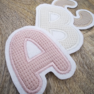 Könnte beinhalten: Drei Filzbuchstaben, A, B und C, in rosa, creme und beige, mit einem weißen Umriss. Die Buchstaben sind mit einem strukturierten Muster bestickt.