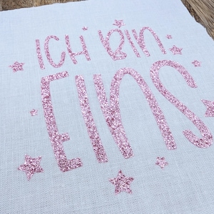 Könnte beinhalten: Ein weißer Stoff mit pinkfarbenem Glitzer-Schriftzug, der "Ich bin eins" lautet, und pinkfarbenen Glitzersternen, die um den Schriftzug verstreut sind.