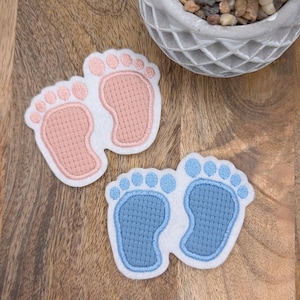 Könnte beinhalten: Zwei Paare gestickter Baby-Fußabdrücke, ein Paar in Rosa und ein Paar in Blau. Die Fußabdrücke befinden sich auf einem weißen Hintergrund.