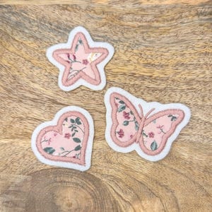 Könnte beinhalten: Drei rosa Stoff-Patches mit Blumendesigns. Die Patches sind in Form eines Sterns, eines Schmetterlings und eines Herzens.