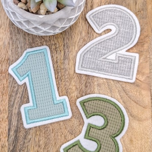 Könnte beinhalten: Drei Filz-Zahlen, eine in hellblau, zwei in grau und drei in grün, mit weißen Umrandungen. Die Zahlen sind in Dreiecksform auf einer Holzoberfläche angeordnet.