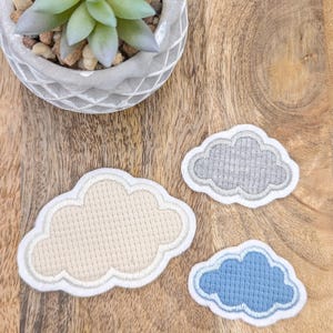 Könnte beinhalten: Drei gestickte Wolkenformen in verschiedenen Farben und Größen. Die Wolken sind beige, grau und blau. Sie befinden sich auf einer Holzoberfläche.