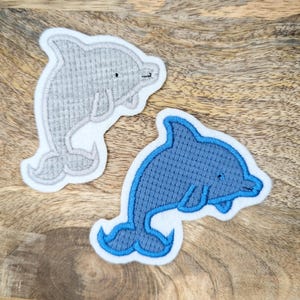Könnte beinhalten: Zwei gestickte Delfin-Aufnäher, einer grau und einer blau, auf einer Holzoberfläche.
