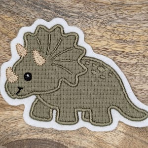 Könnte beinhalten: Ein gestickter Aufnäher mit einem Cartoon-Triceratops-Dinosaurier, grün mit weißem Rand.
