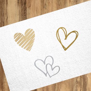 Könnte beinhalten: Drei handgezeichnete Herzen auf weißem Hintergrund. Ein Herz ist goldfarben mit einem gestreiften Muster, ein anderes ist goldfarben mit einem einfachen Umriss und das letzte ist ein Paar grauer Herzen.