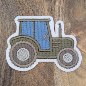 Könnte beinhalten: Gestickter Aufnäher eines grünen und blauen Traktors mit weißem Rand. Der Traktor hat große Räder, eine Kabine mit Lenkrad und einen Schornstein. Der Aufnäher ist zum Aufbügeln bestimmt.