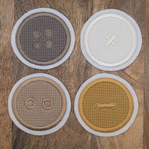 Könnte beinhalten: Vier runde Untersetzer mit Knopfdesigns. Jeder Untersetzer hat eine andere Farbe: Grau, Off-White, Beige und Senfgelb. Die Knopfformen sind in passenden Farben mit einem weißen Rand gestickt.