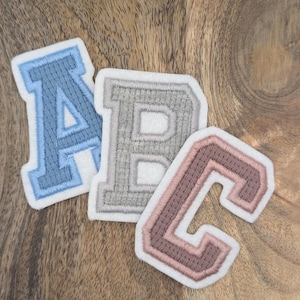 Könnte beinhalten: Drei gestickte Stoffaufnäher in Form der Buchstaben A, B und C. Der Buchstabe A ist hellblau, B ist grau und C ist rosa. Jeder Buchstabe hat einen weißen Rand, vor einem Holzgrund.