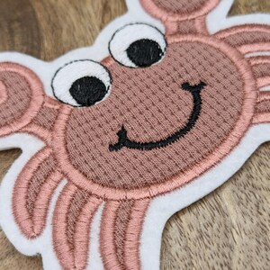 Könnte beinhalten: Eine gestickte rosa Krabbe mit weißen Augen und einem schwarzen Lächeln. Die Krabbe befindet sich auf einem weißen Hintergrund.