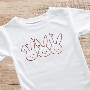 Könnte beinhalten: Weißes T-Shirt mit einer braunen Umrisszeichnung von drei Cartoon-Hasen, jeweils mit einem Herz. Die Hasen haben einfache Gesichter und sind in einer Reihe angeordnet. Ein charmantes und verspieltes Design.