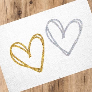 Könnte beinhalten: Zwei Glitzerherzen, eines golden und eines silbern, auf weißem Hintergrund.