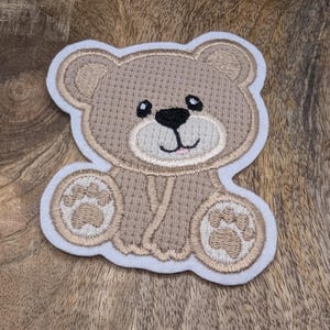 Könnte beinhalten: Ein beigefarbener gestickter Teddybär-Patch mit weißem Rand. Der Bär hat ein lächelndes Gesicht mit schwarzen Augen und einer schwarzen Nase.