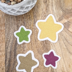 Könnte beinhalten: Vier gestickte Filzsterne in verschiedenen Farben: Gelb, Grün, Braun und Lila. Die Sterne liegen auf einer Holzoberfläche.