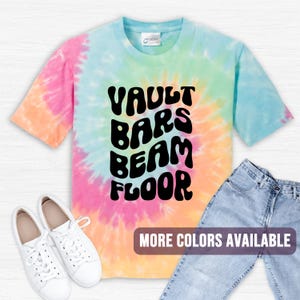 May include: A tie-dye t-shirt with the words "VAULT BARS BEAM FLOOR" in black. The shirt is a mix of pink, yellow, green, and blue. White sneakers and blue jeans are next to the shirt. Text at the bottom says "MORE COLORS AVAILABLE."