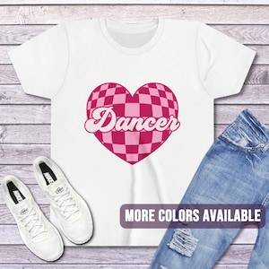May include: White t-shirt with a pink checkered heart and the word "Dancer" in a retro font. The shirt is laying flat on a wooden surface with a pair of white sneakers and blue jeans.