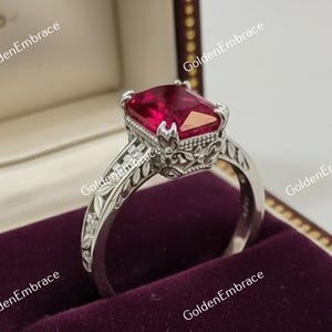 May include: A silver ring with a large, rectangular red gemstone, set in an ornate, floral-patterned band. The ring is displayed in a maroon velvet-lined jewelry box with a gold clasp and a brown wooden exterior.