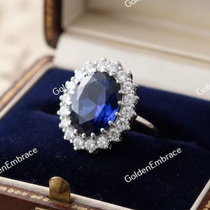 May include: A silver ring featuring a large, oval, deep blue gemstone encircled by a halo of smaller, clear stones. The ring is presented in a dark brown, hinged jewellery box with a blue velvet interior.