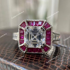 May include: A silver ring with a large, square, clear center stone, surrounded by smaller, square, red stones. The ring has a detailed band with additional small, clear stones. The ring is set on a textured, brown surface.