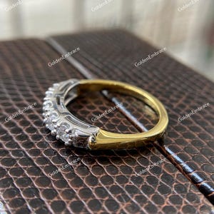 May include: A gold and silver band ring with a row of small, clear gemstones. The ring is on a brown, textured surface.