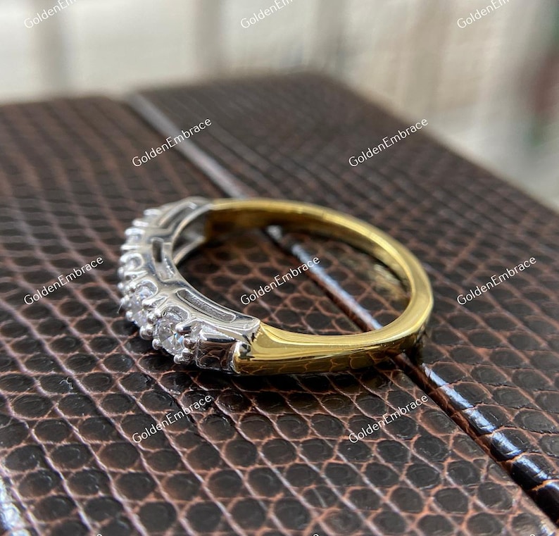 May include: A gold and silver band ring with a row of small, clear gemstones. The ring is on a brown, textured surface.