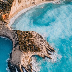 May include: Aerial view of Porto Katsiki beach in Greece. Turquoise water surrounds a rocky cliff covered in green vegetation. Beige cliffs and a sandy beach are visible in the background. The image is taken from a high angle.