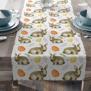 Könnte beinhalten: Weißer Tischläufer mit einem sich wiederholenden Muster aus braunen Hasen, gelben und orangefarbenen Ostereiern und grünen und violetten Blütenstielen.