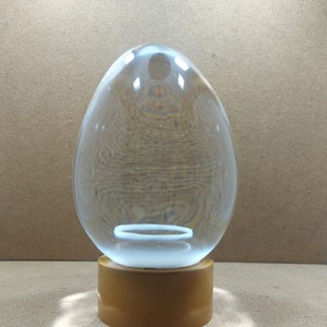 Pode incluir: Um ornamento de vidro transparente em forma de ovo com uma base dourada. O ornamento est&aacute; sobre uma superf&iacute;cie marrom.