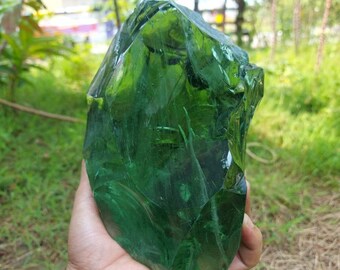 May include: A large, green, irregularly shaped piece of glass, likely a piece of sea glass, is held in a person's hand. The glass is translucent and has a smooth, polished surface.