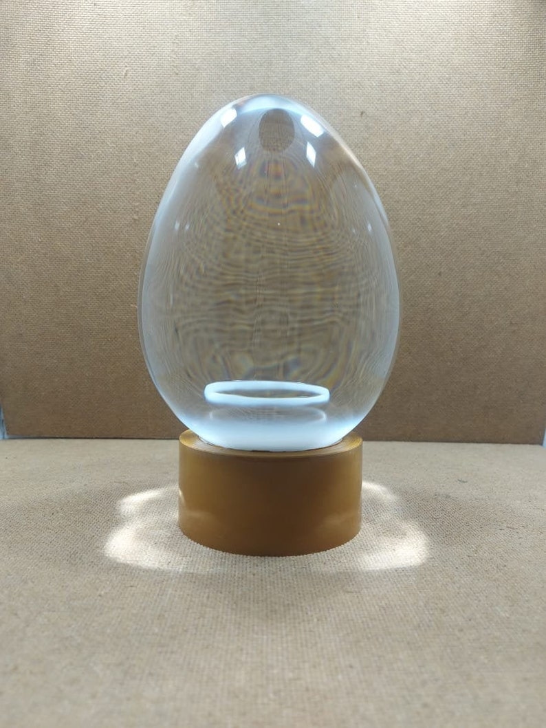 Pode incluir: Um ornamento de vidro transparente em forma de ovo com uma base dourada. O ornamento est&aacute; sobre uma superf&iacute;cie marrom.