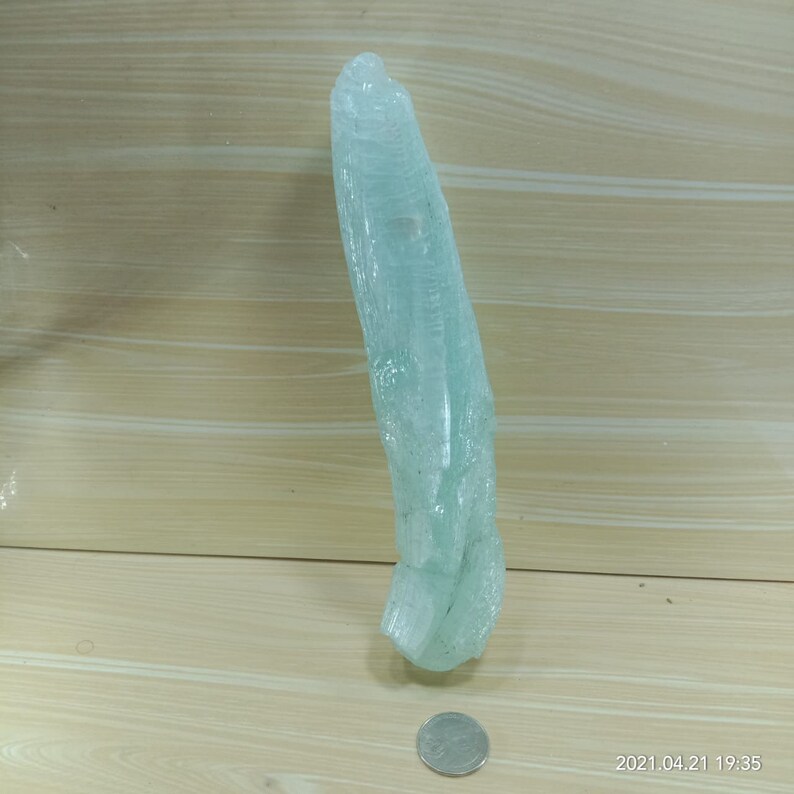 May include: A large, light blue crystal with a pointed tip and a rough, textured surface. The crystal is about 15 inches long and is sitting on a wooden surface.